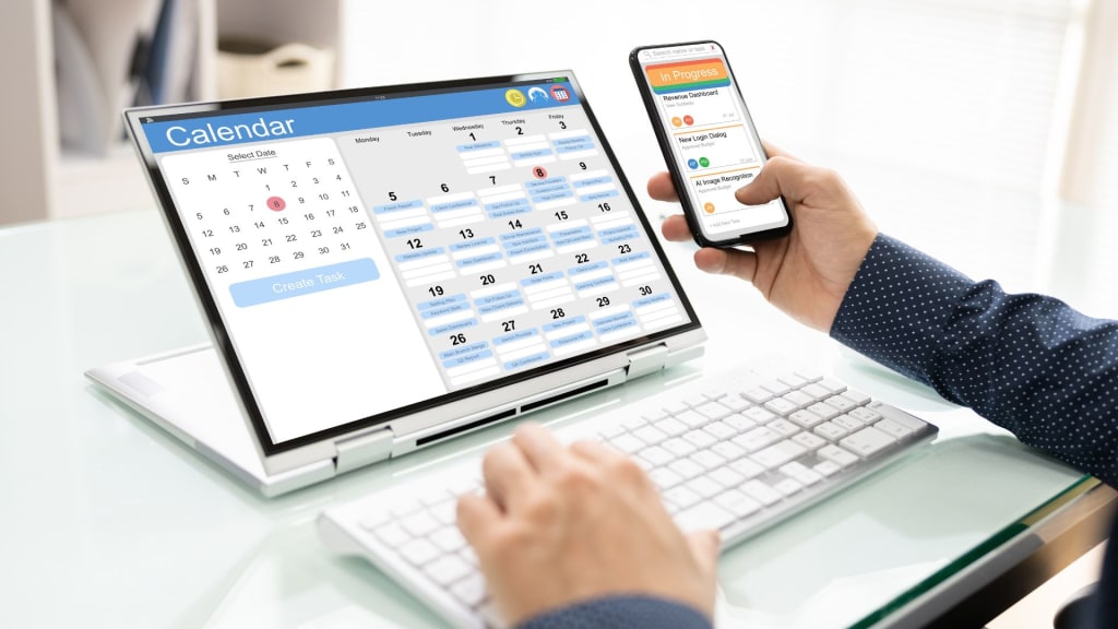 Person working at a desk, viewing a digital calendar on a laptop while holding a smartphone displaying therapist scheduling software for easy appointment and task management.